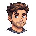 Young man with tousled brown hair, a light beard, and a gentle smile. He has large grey and blue expressive eyes and wears a grey t-shirt.