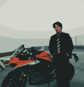A person in a dark suit stands next to a black and orange sporty motorcycle.