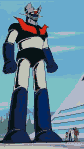 Colossal robot with dark blue, white, and red armor, a red V-emblem on its chest, and a metallic head, stands before a futuristic building. Smaller figures and vehicles are visible near its legs.